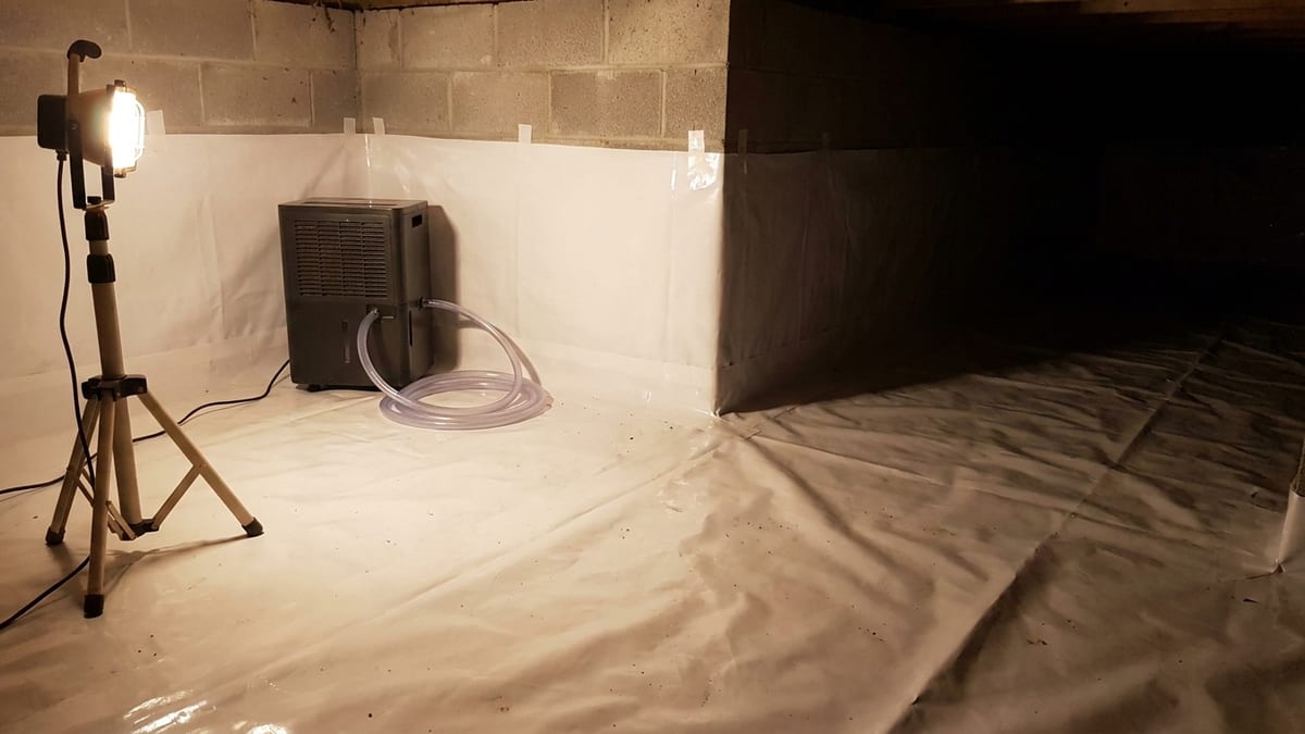 Interior of a residential crawl space with bright white vapor barrier sheeting laid over the soil, taped seams running up a block foundation wall, and a dehumidifier with a coiled drain line in the corner under a work light