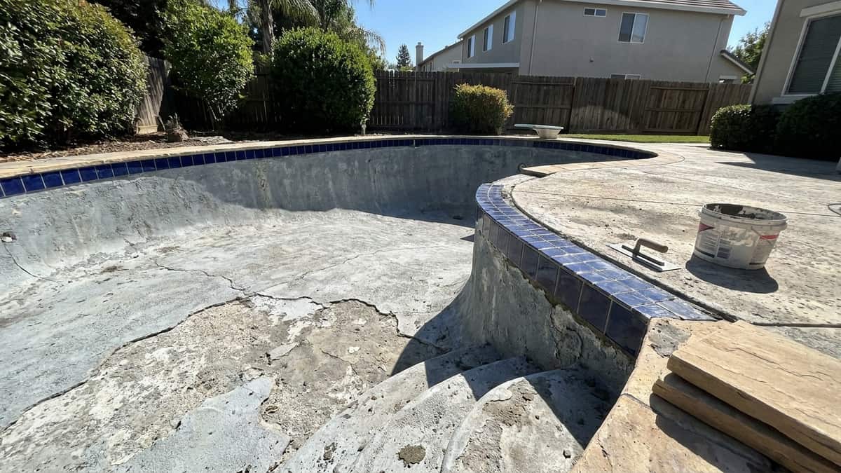 Drained residential swimming pool mid-resurfacing with worn plaster chipped out of the shell, a new tile band being set along the waterline, mortar bucket and trowel on the deck, and coping stones stacked nearby in bright midday light